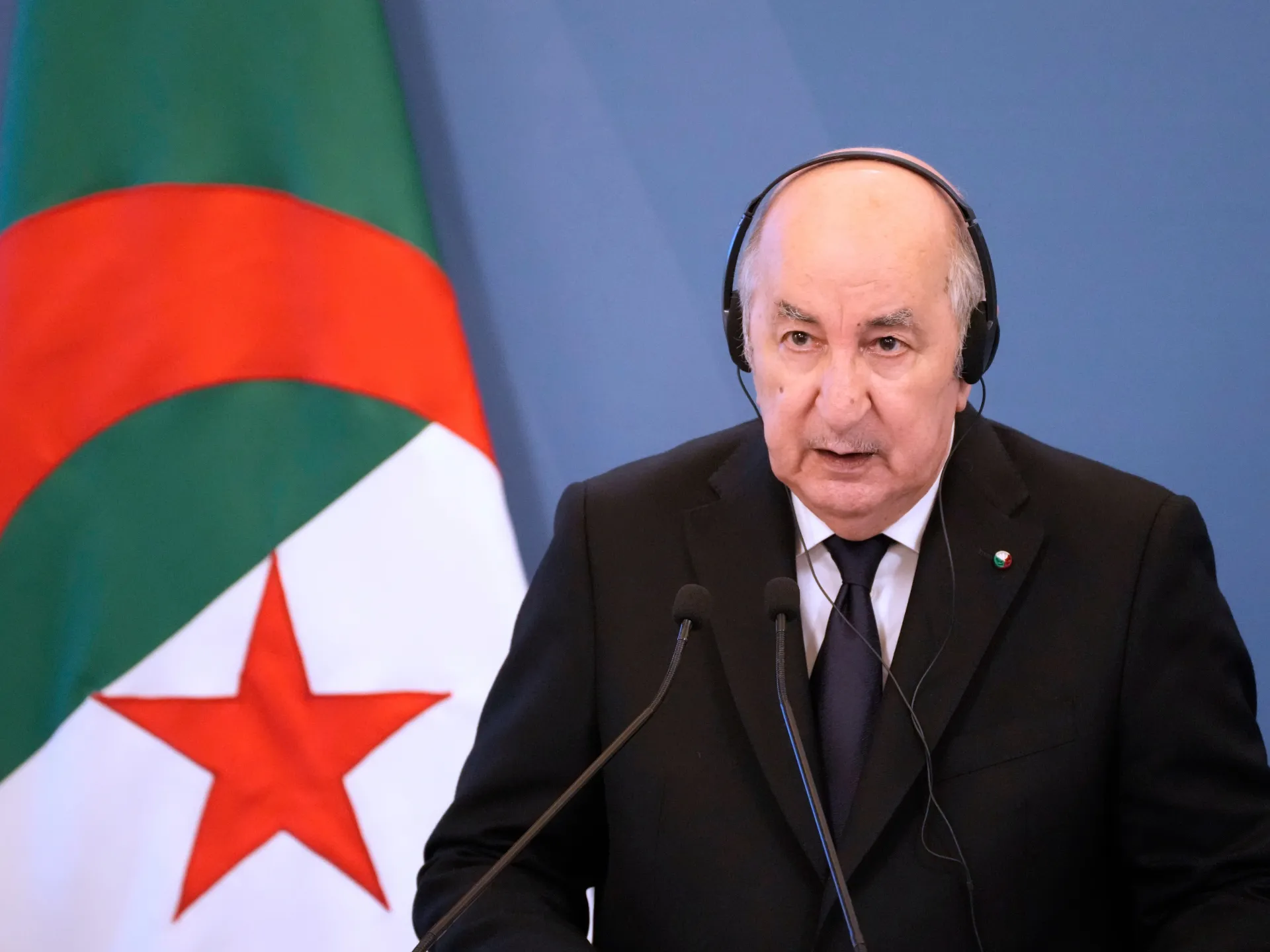 Man in a suit and headphones speaks at microphones beside an Algerian flag.