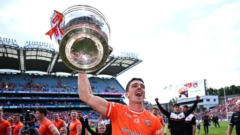 An orange-shirted player raises a large silver trophy above his head on a crowded pitch.