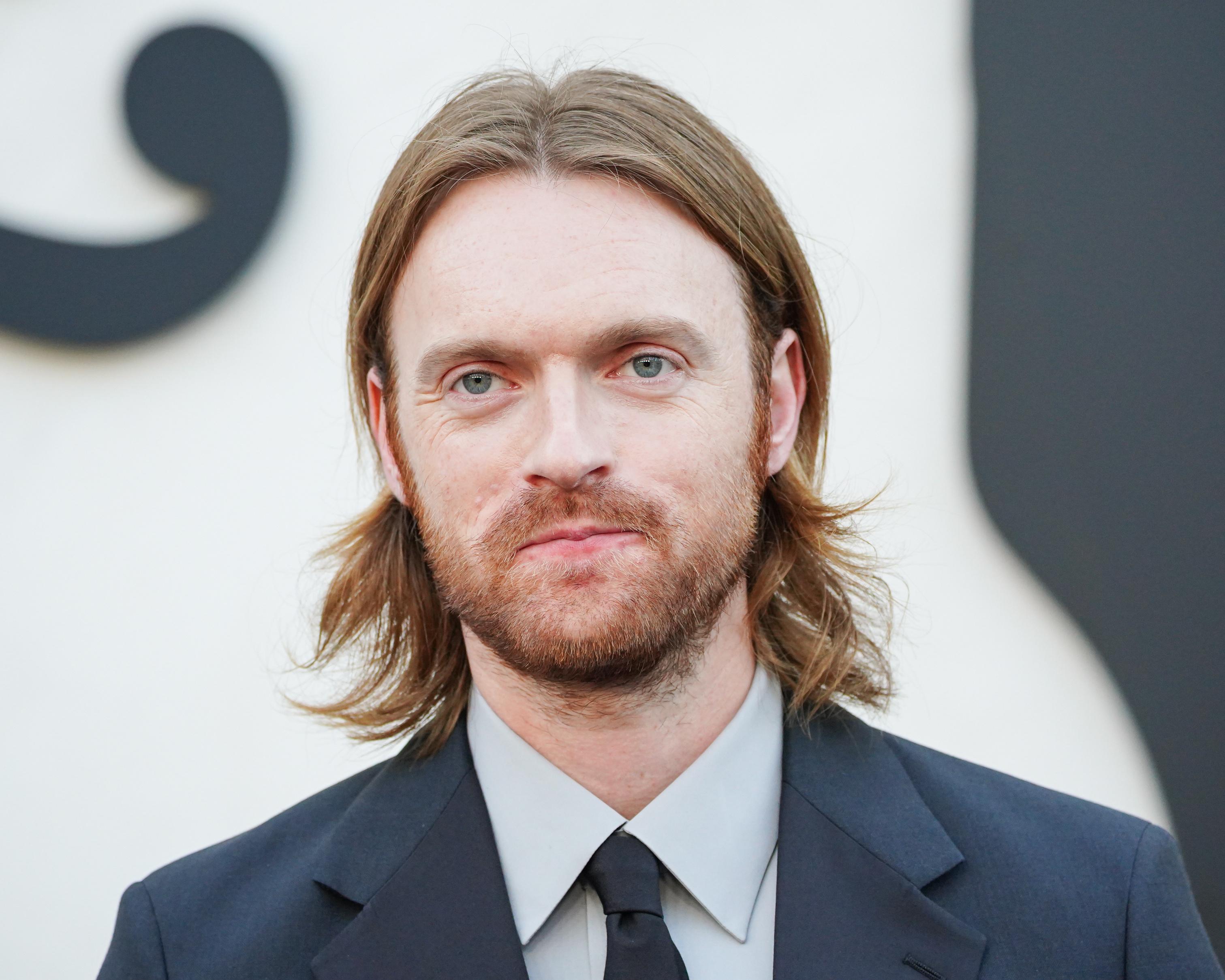 Close-up portrait of a long-haired, bearded man wearing a dark suit and tie.