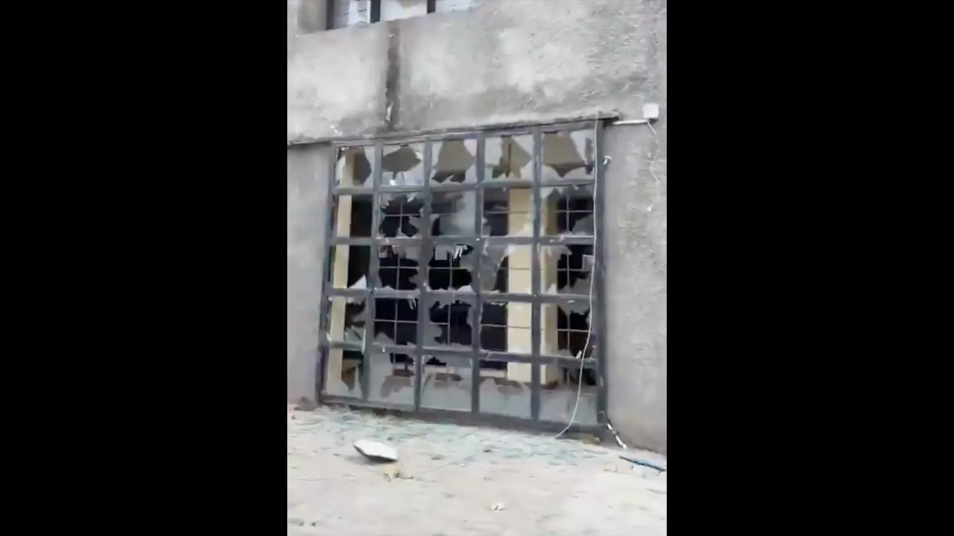 A damaged metal-framed glass entrance with shattered panes and debris scattered on the ground.