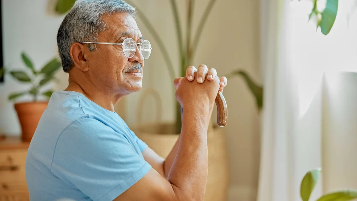 Older man in glasses sits indoors, clasping a curved cane near a sunlit window.