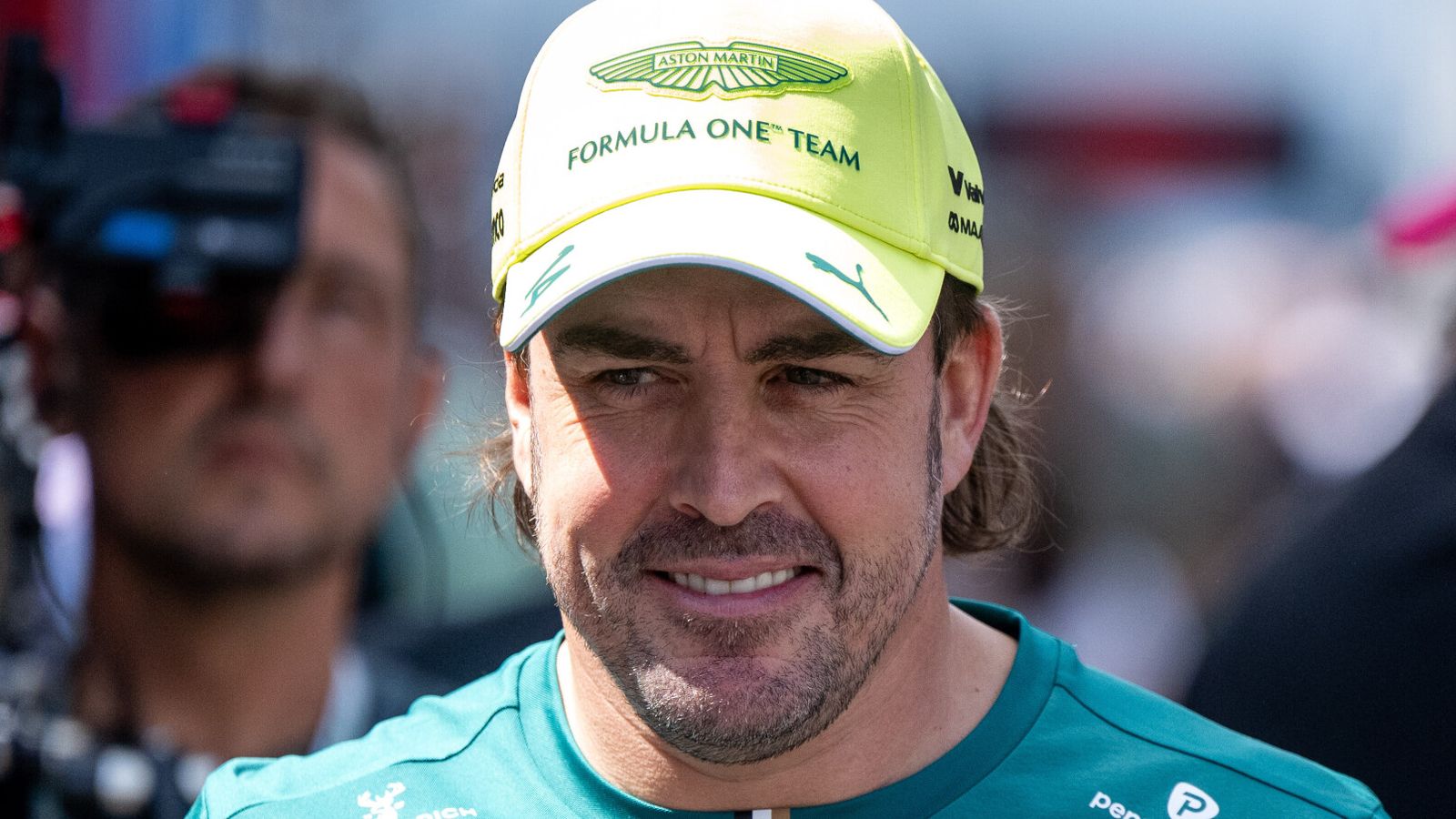 Fernando Alonso smiles in an Aston Martin cap and teal team shirt at a racing event.