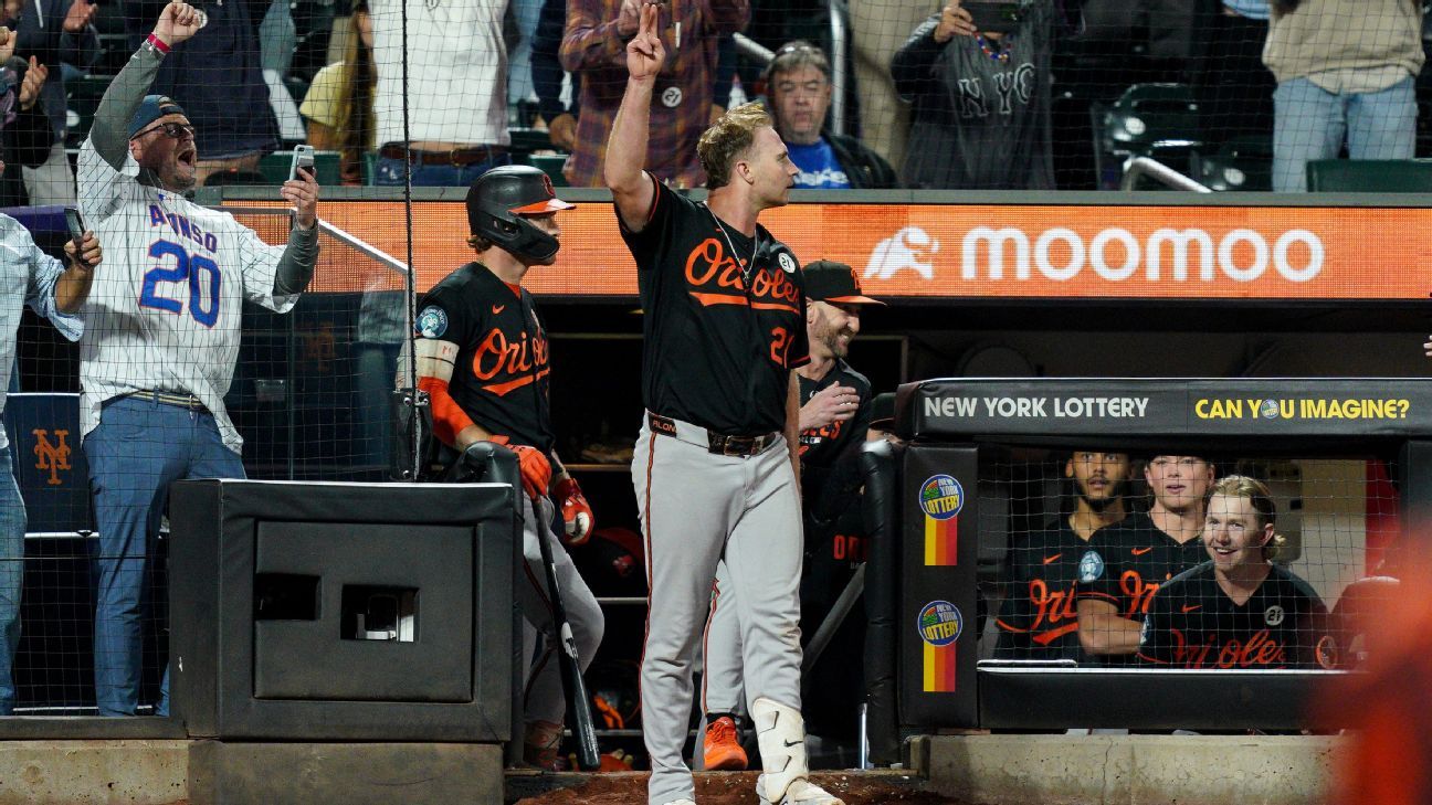 Baseball player raises three fingers near dugout as teammates and fans celebrate behind him