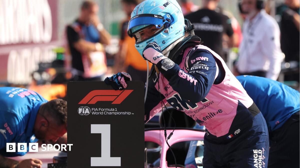 Helmeted Formula 1 driver celebrates beside a large number one podium board.