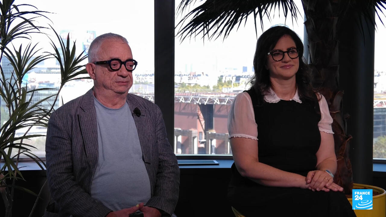 A man and a woman wearing glasses stand indoors in front of large windows and plants.