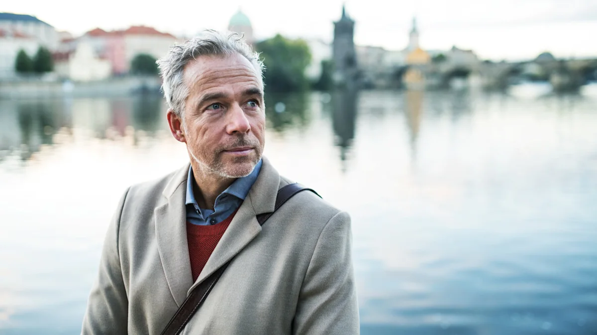 Gray-haired man in coat standing beside a waterfront with blurred city buildings behind him