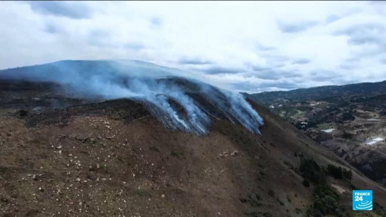 Smoke rises from a barren mountainside above a valley settlement under cloudy skies.
