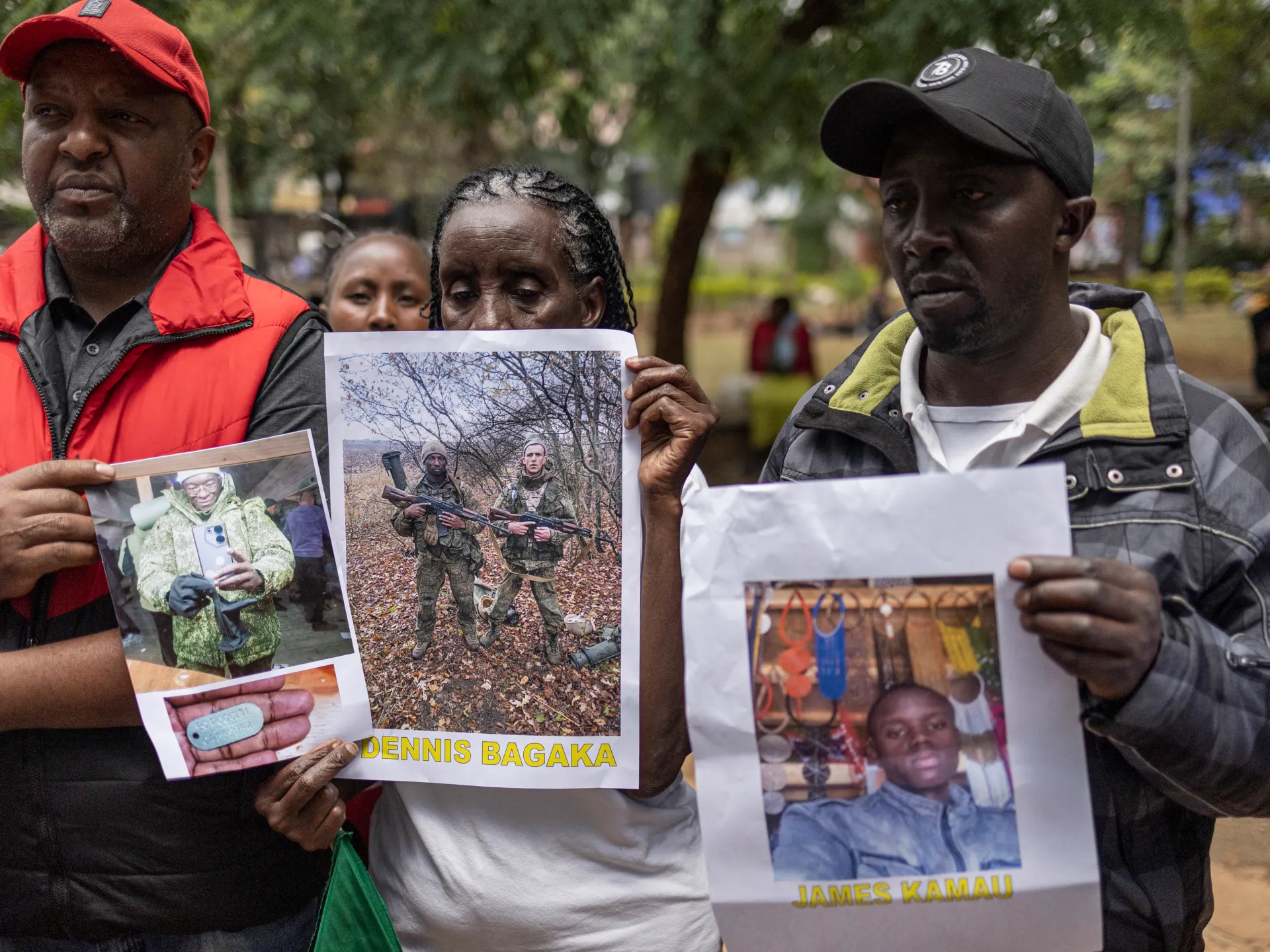 Three people hold posters showing photographs of men, including a soldier and two civilians.