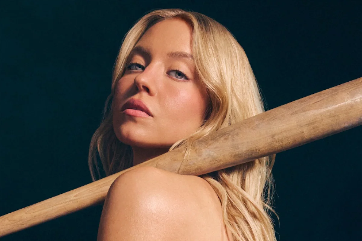 Blonde woman posing with a wooden baseball bat resting across her shoulders against a dark background