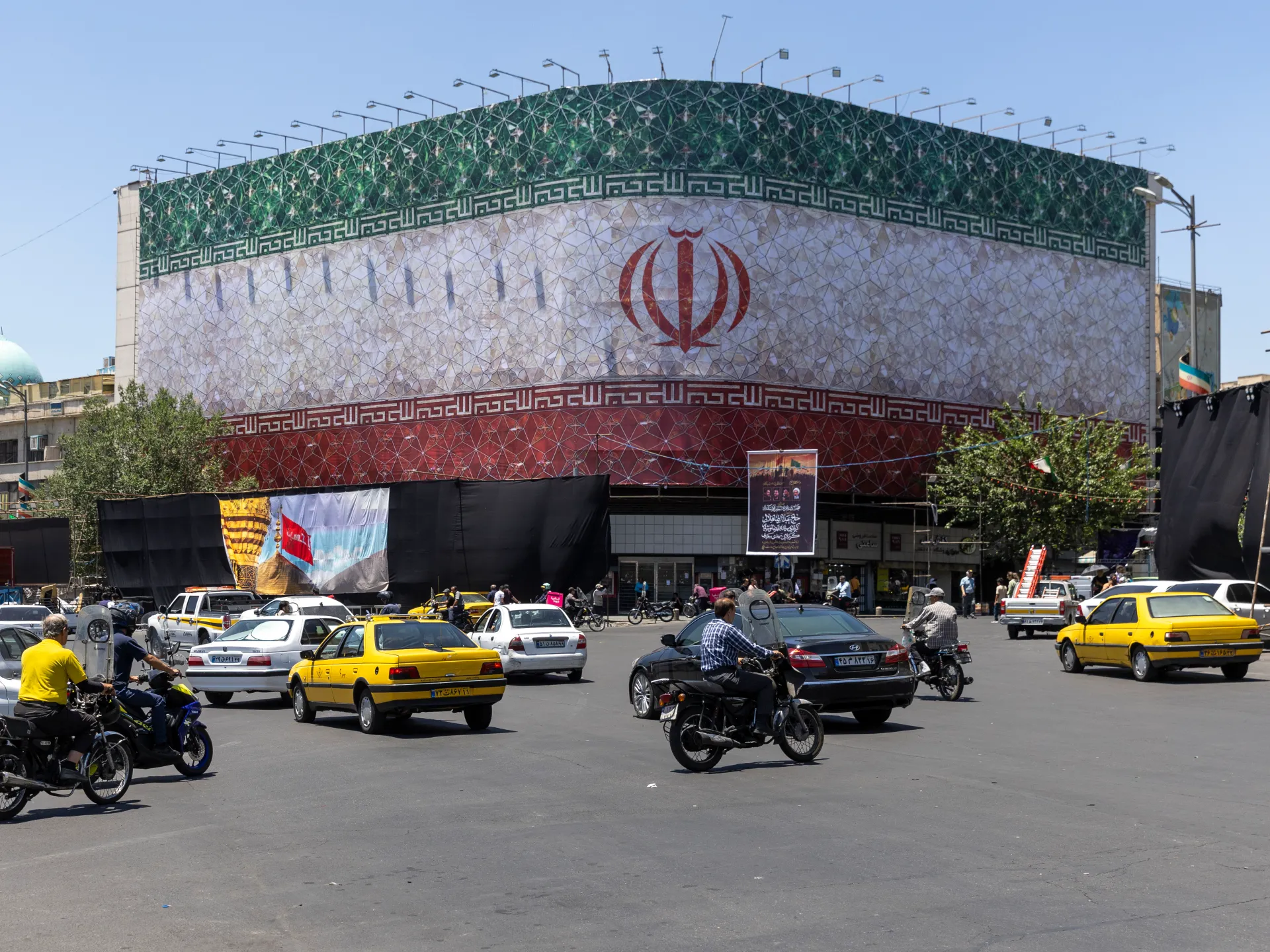 Yellow taxis, motorcycles, and cars fill a busy Iranian city intersection before a decorated building.