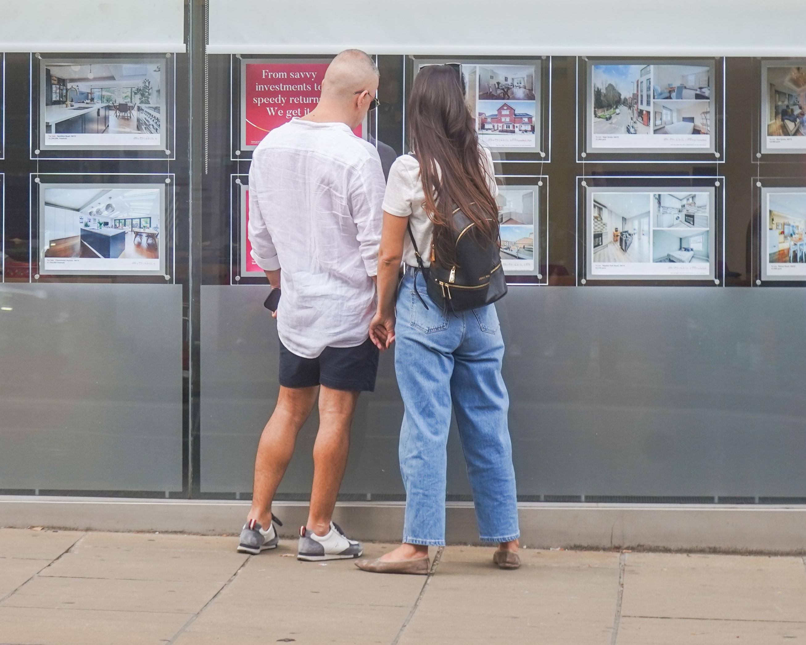 A couple look at property listings displayed in a real estate office window.