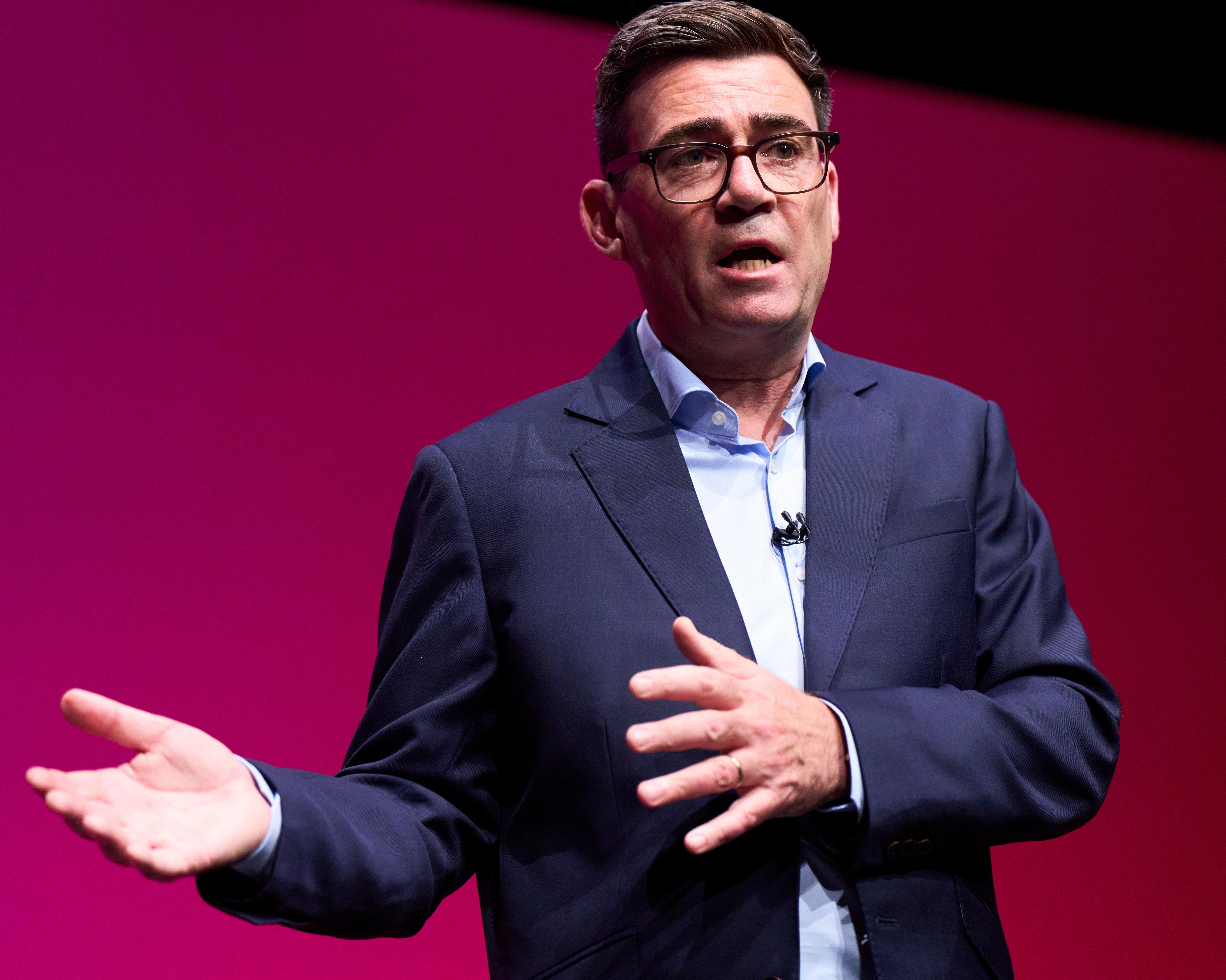 A bespectacled man in a navy suit gestures while speaking against a magenta stage backdrop.