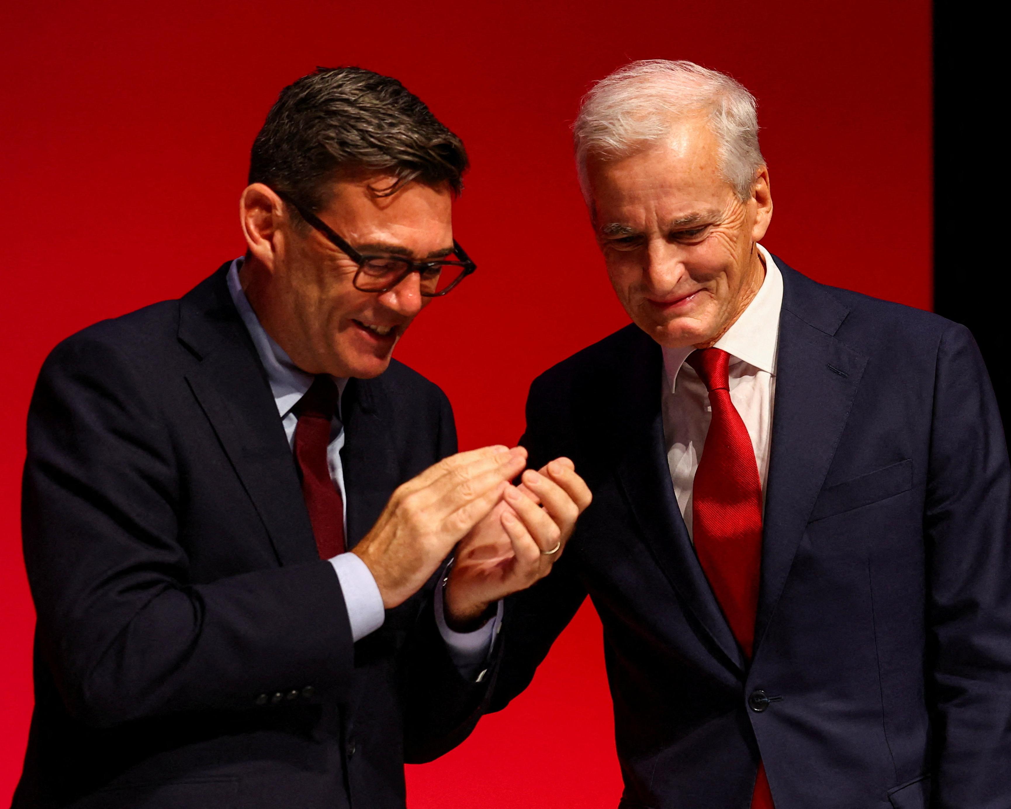 Two men in dark suits look down at their joined hands against a red backdrop.