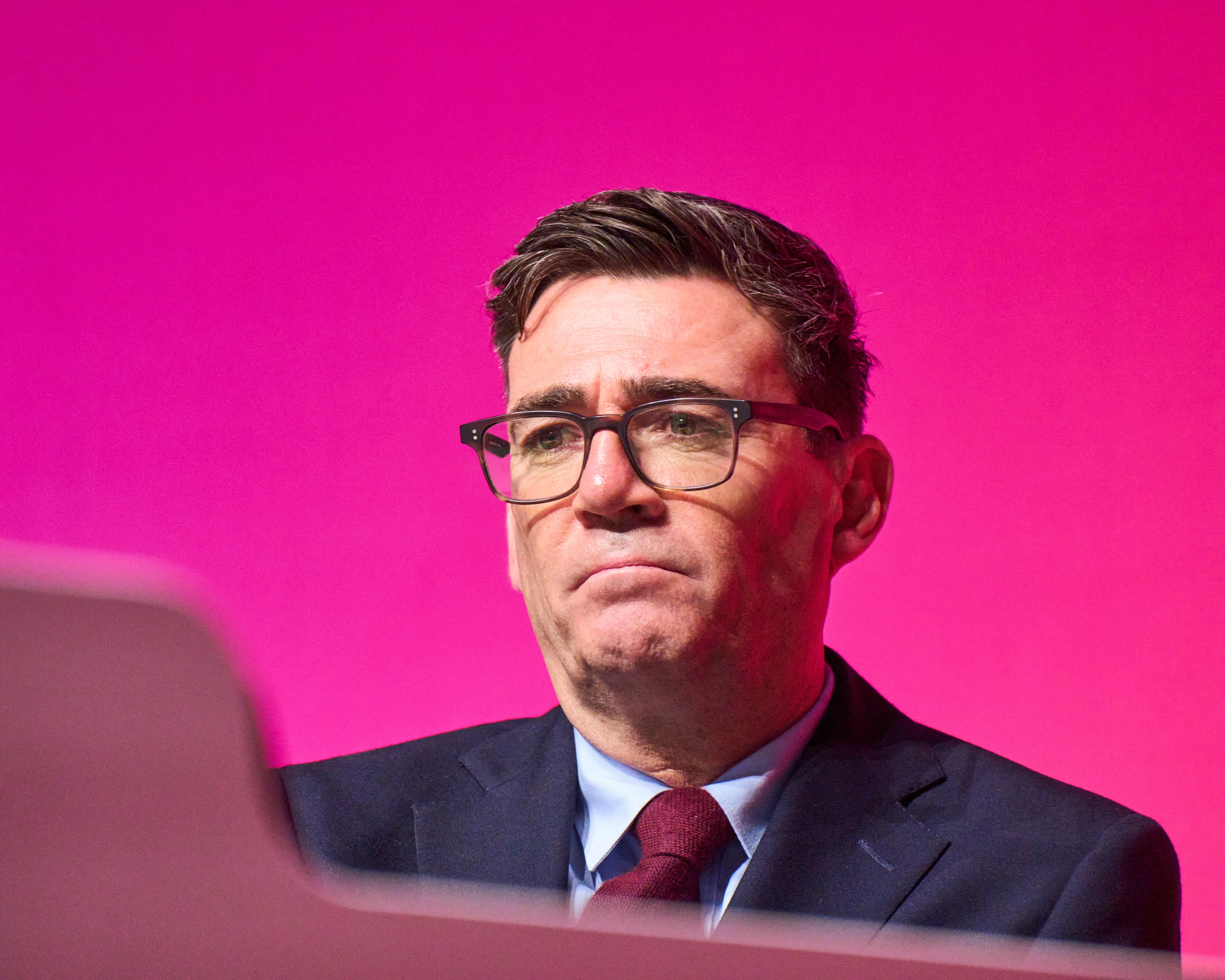 A bespectacled man in a dark suit and red tie stands against a bright pink backdrop.