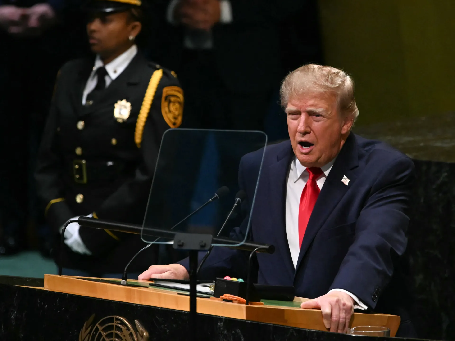 Donald Trump speaks at a lectern with microphones, with a uniformed officer standing behind him.