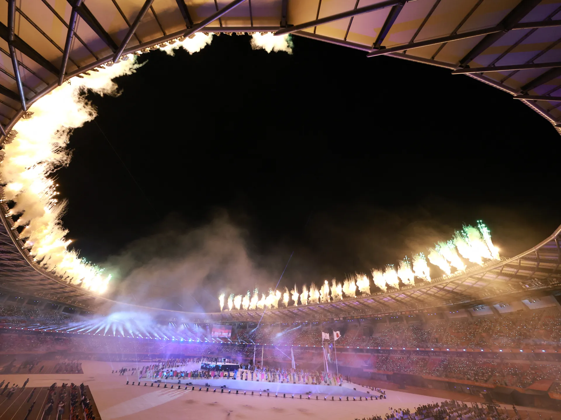 Fireworks erupt above a stadium during a brightly lit nighttime ceremony with performers on the field.