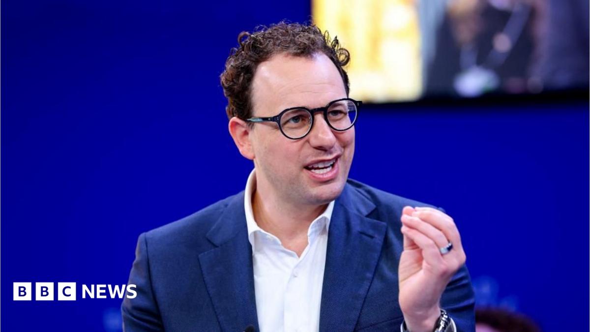 Man wearing glasses and a dark suit speaks while gesturing in front of a blue BBC News backdrop.