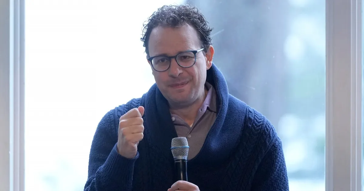 A bespectacled man in a navy cardigan holds a microphone and raises a fist near a bright window.