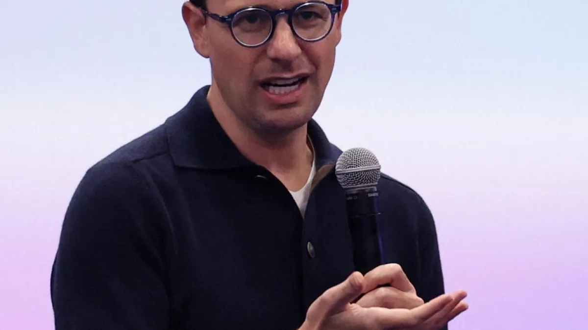 Man wearing round glasses speaks into a handheld microphone against a pale background.