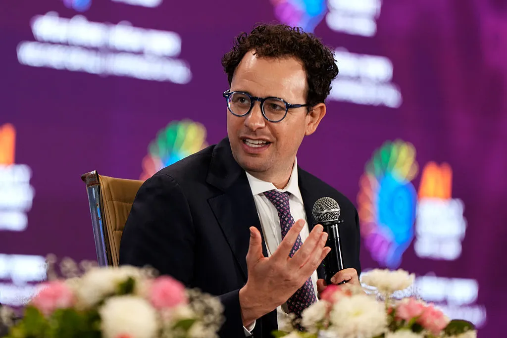 Man in suit and glasses speaking into a microphone during a panel discussion with floral arrangements foregrounded
