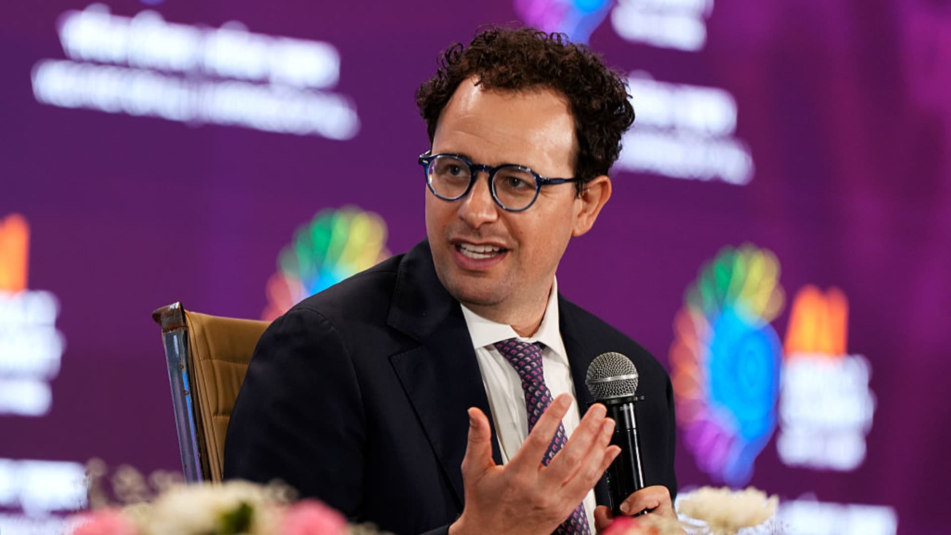 Man in glasses and suit speaking into a microphone during a stage discussion