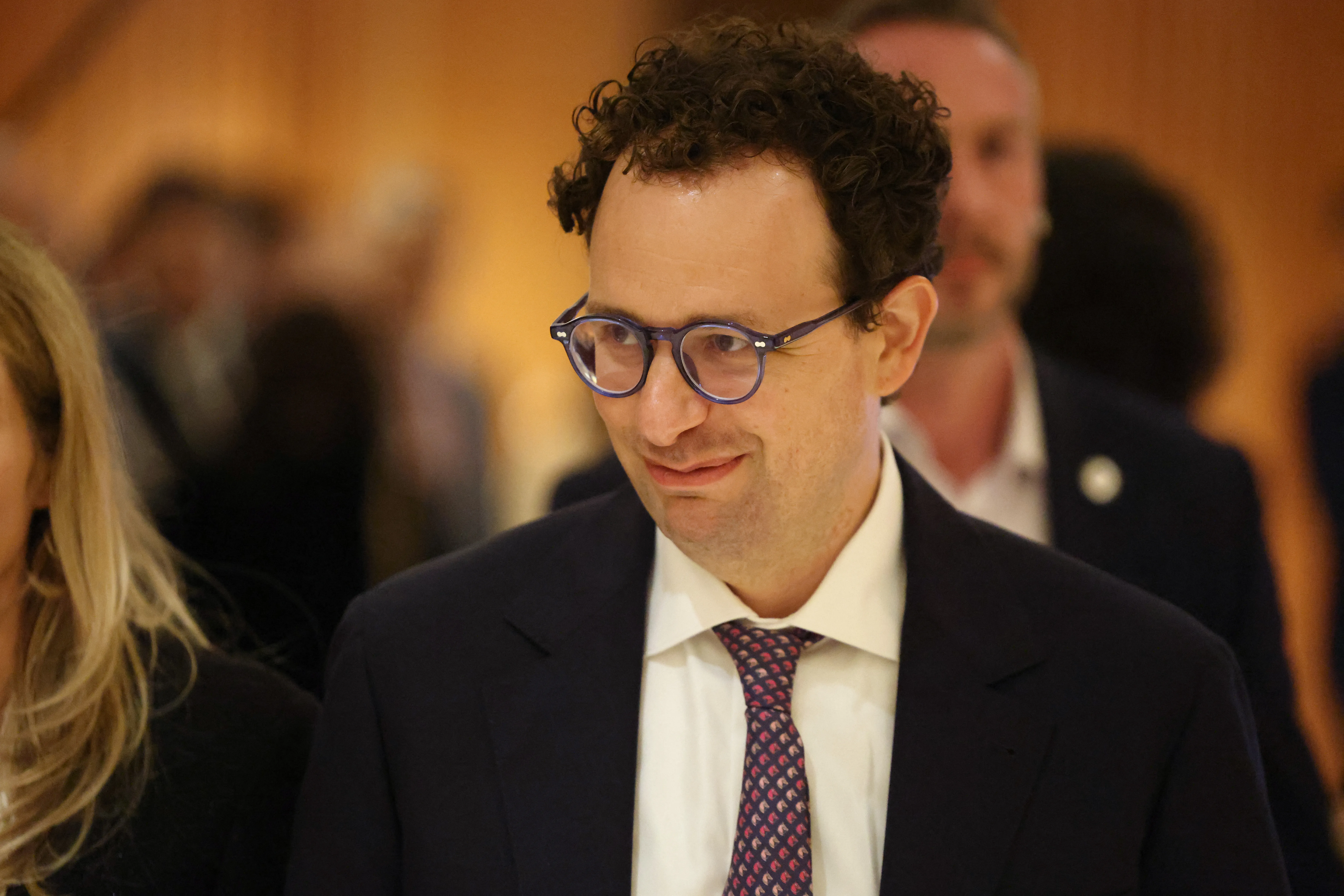 A curly-haired man in glasses, a dark suit, and patterned tie stands among formally dressed guests.