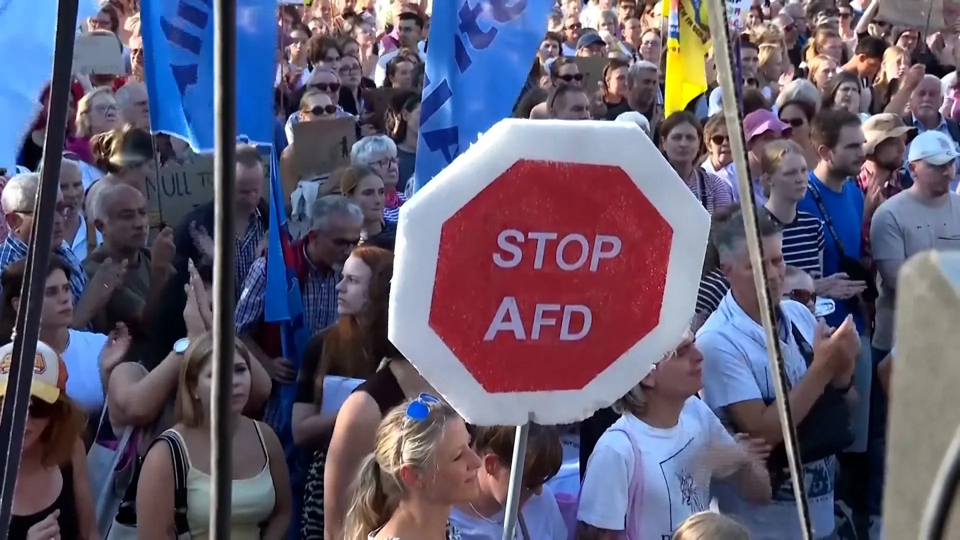 Crowd of protesters holding blue flags behind a large red-and-white STOP AfD sign