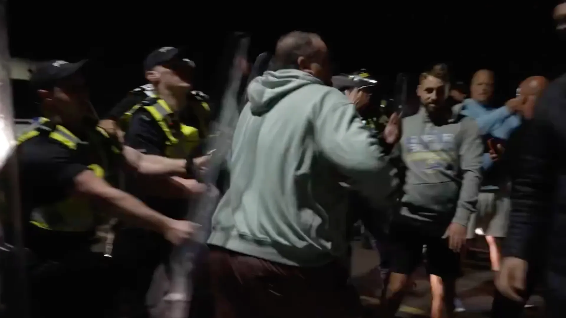 Police officers restrain a man in a gray hoodie amid a nighttime crowd.