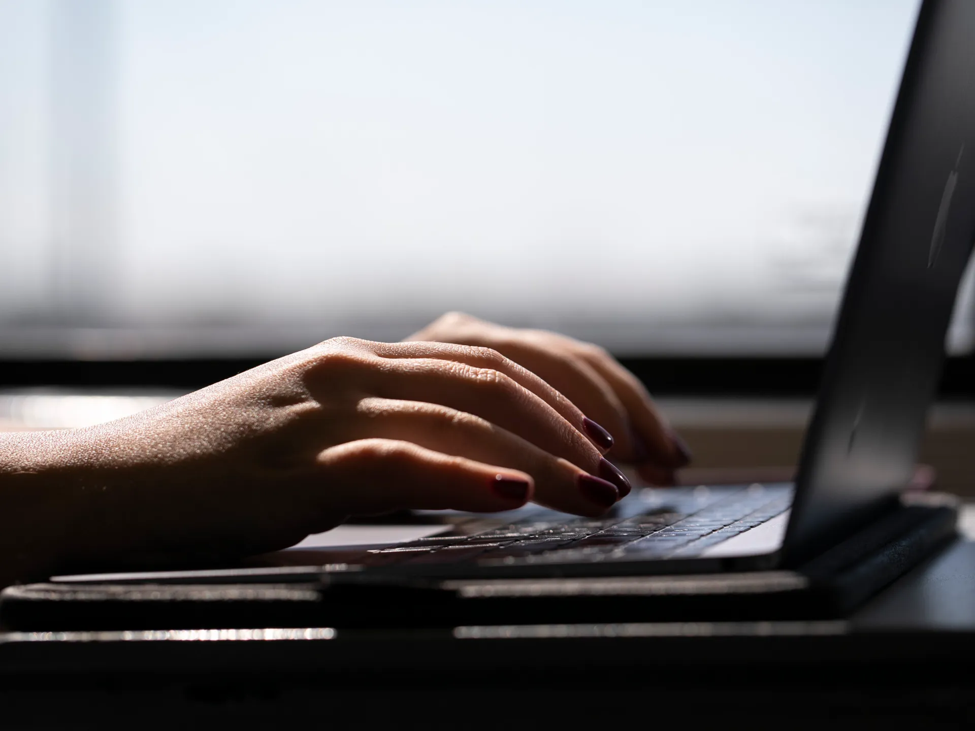 Close-up of hands with dark nail polish typing on a laptop keyboard in low light.