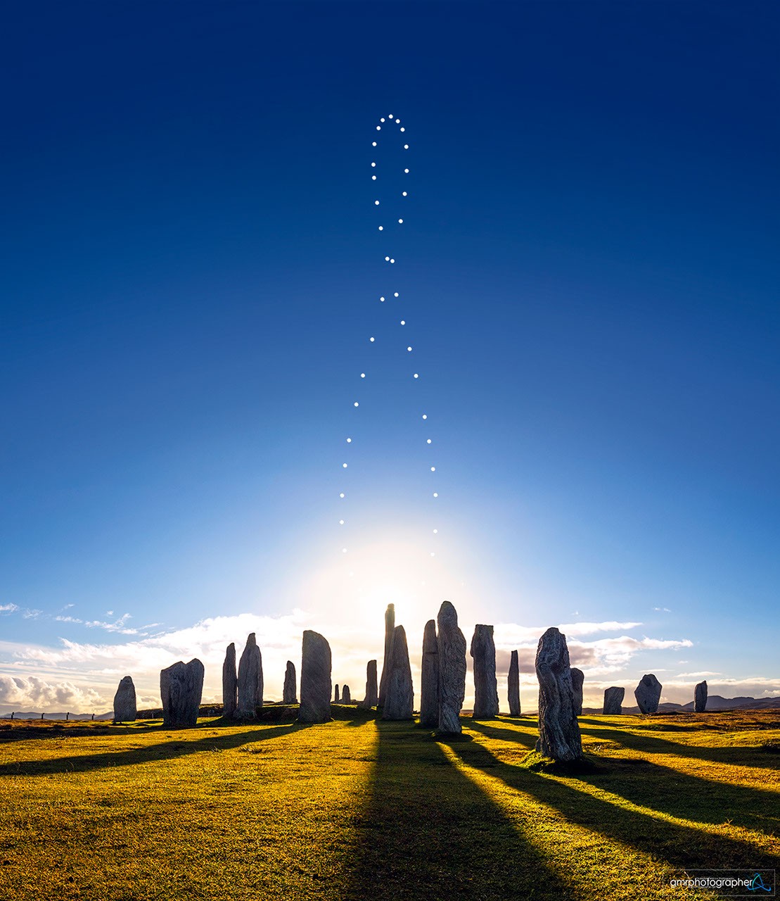 Stone circle standing in a sunlit grassy landscape beneath a blue sky with dotted analemma