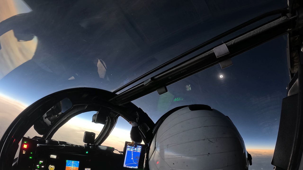 View from an aircraft cockpit showing a helmeted pilot, instruments, windshield, and cloud-lined horizon