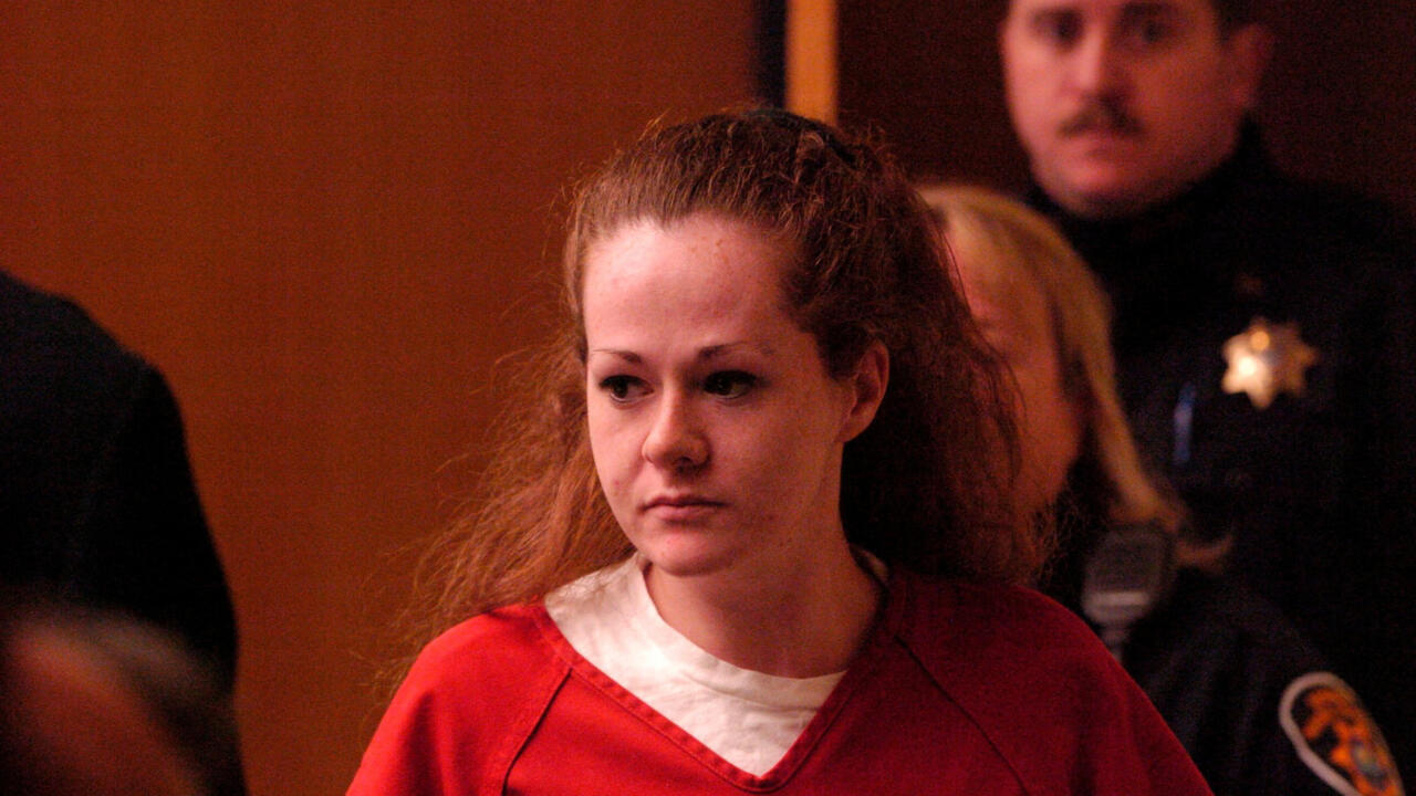A woman in a red top sits indoors, with several people visible behind her.