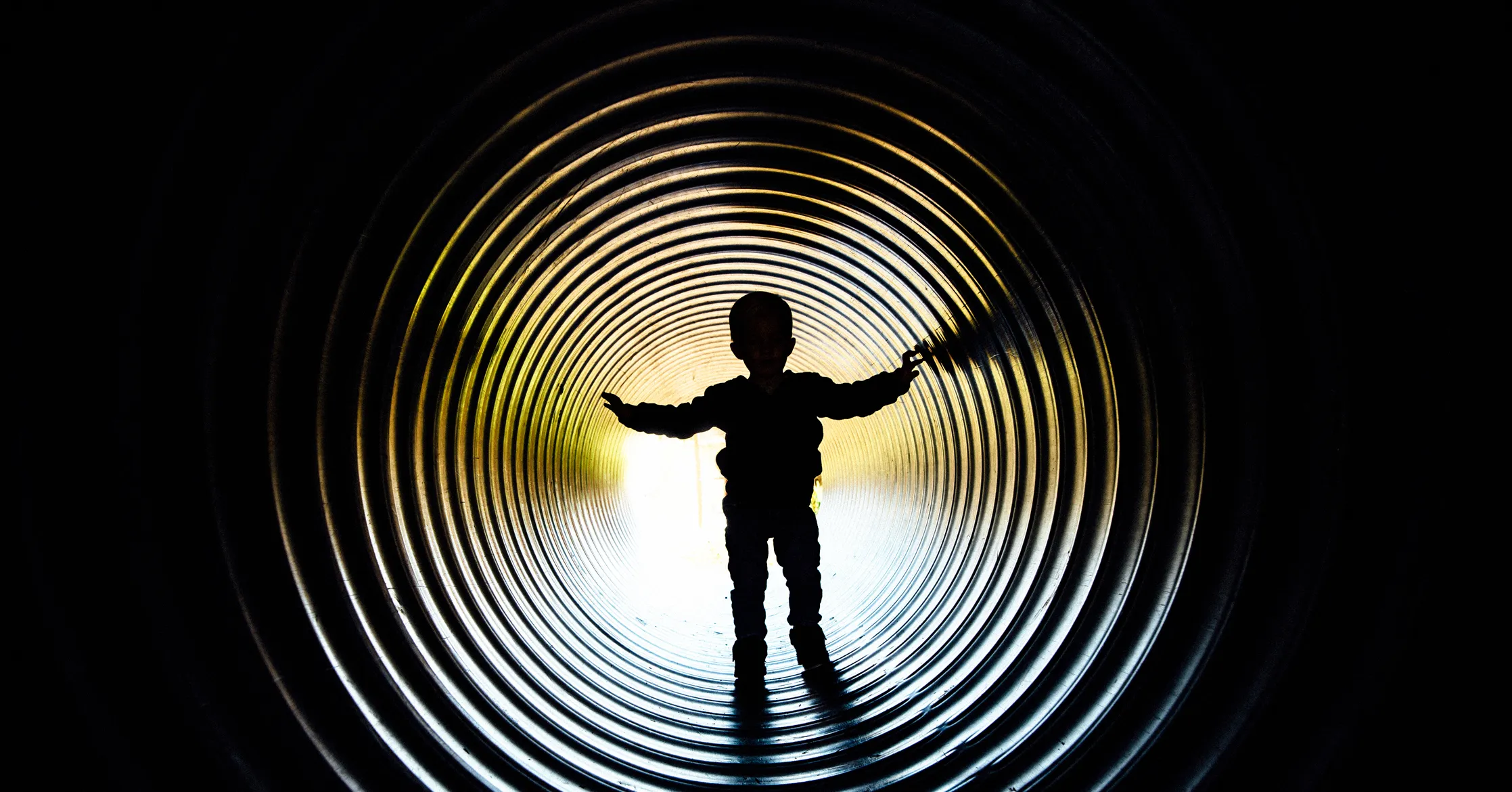 Silhouetted child stands inside a brightly lit corrugated tunnel, arms outstretched.