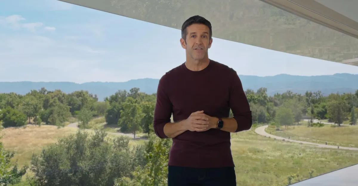 Man in burgundy sweater stands indoors before large windows overlooking a green landscape.