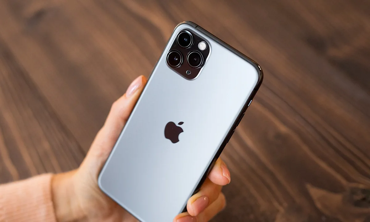 Hand holding a silver iPhone with three rear cameras over a wooden surface