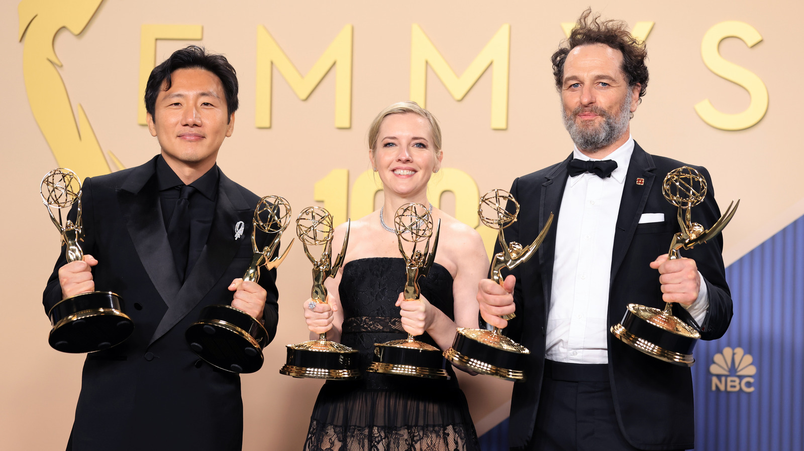 Three formally dressed people pose together, holding multiple Emmy statuettes against an Emmys backdrop.