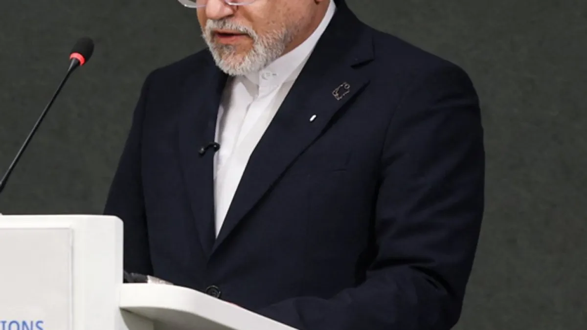 Bearded man in a dark suit speaks at a white lectern beside a microphone.