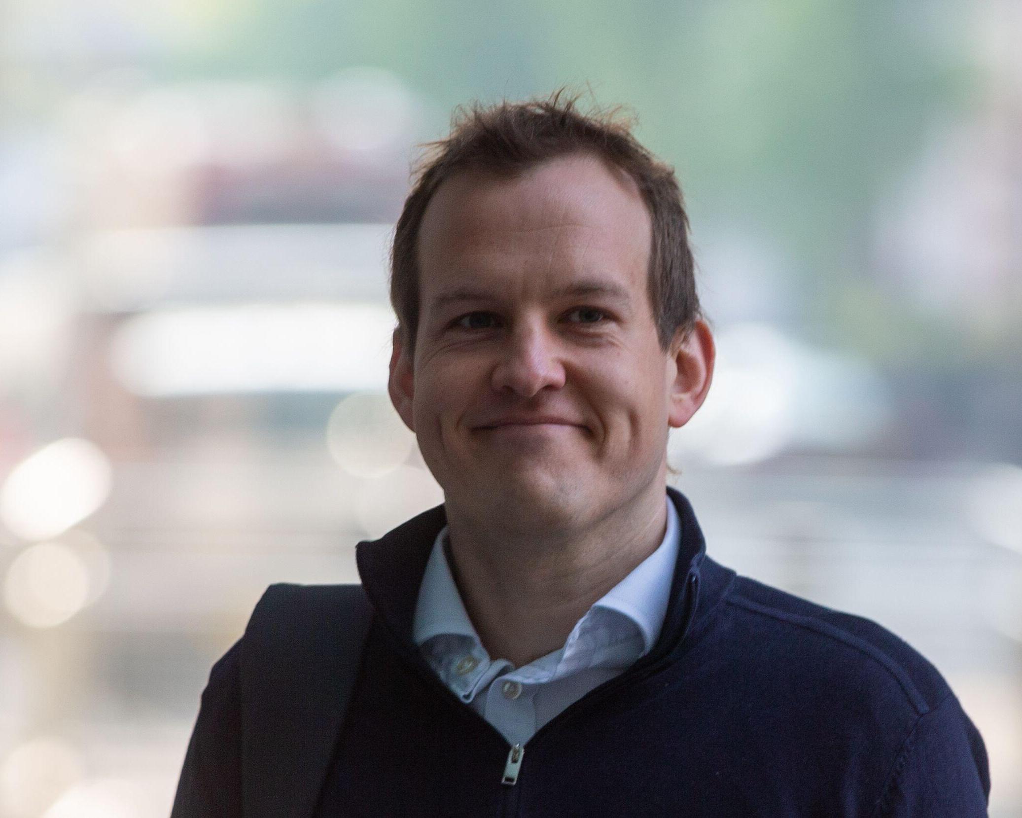Smiling man wearing a dark jacket and collared shirt in a softly blurred indoor setting