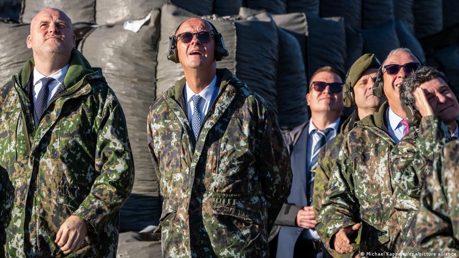 Men in camouflage jackets and formal attire look upward during an outdoor gathering.