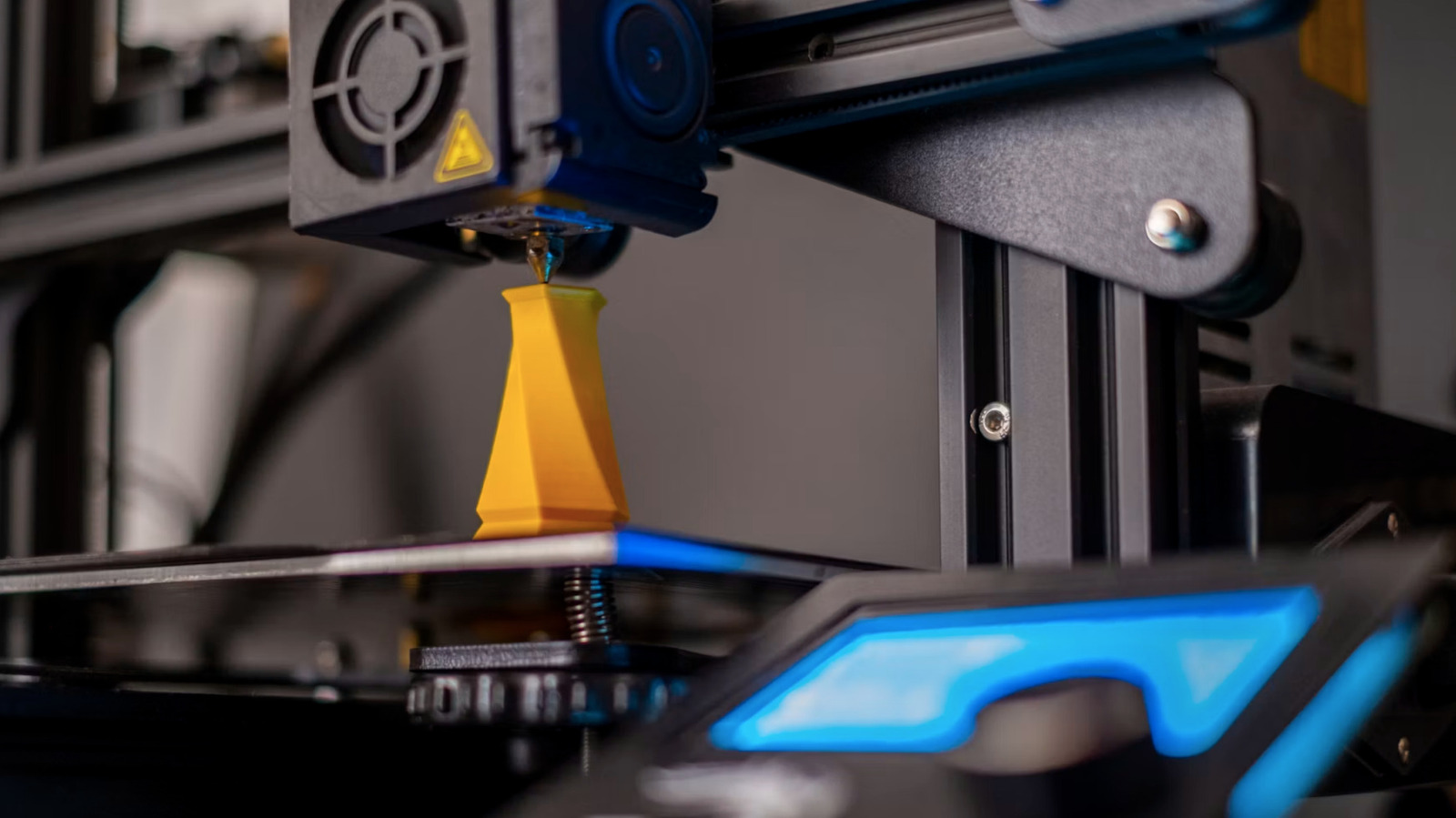 A 3D printer deposits yellow filament onto an object, with blue lights glowing on its frame.