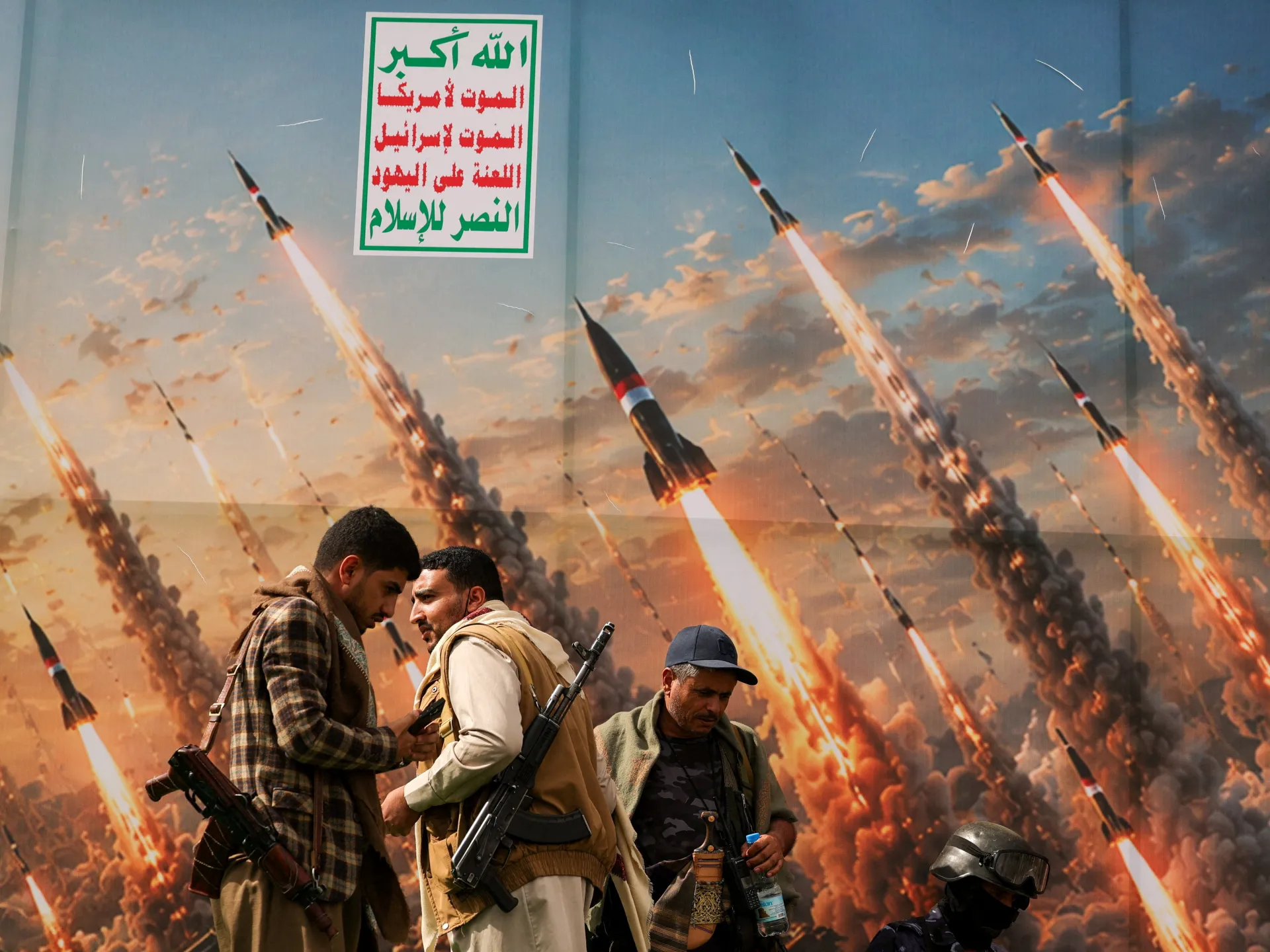 Armed men stand before a mural depicting missiles launching beneath an Arabic slogan.