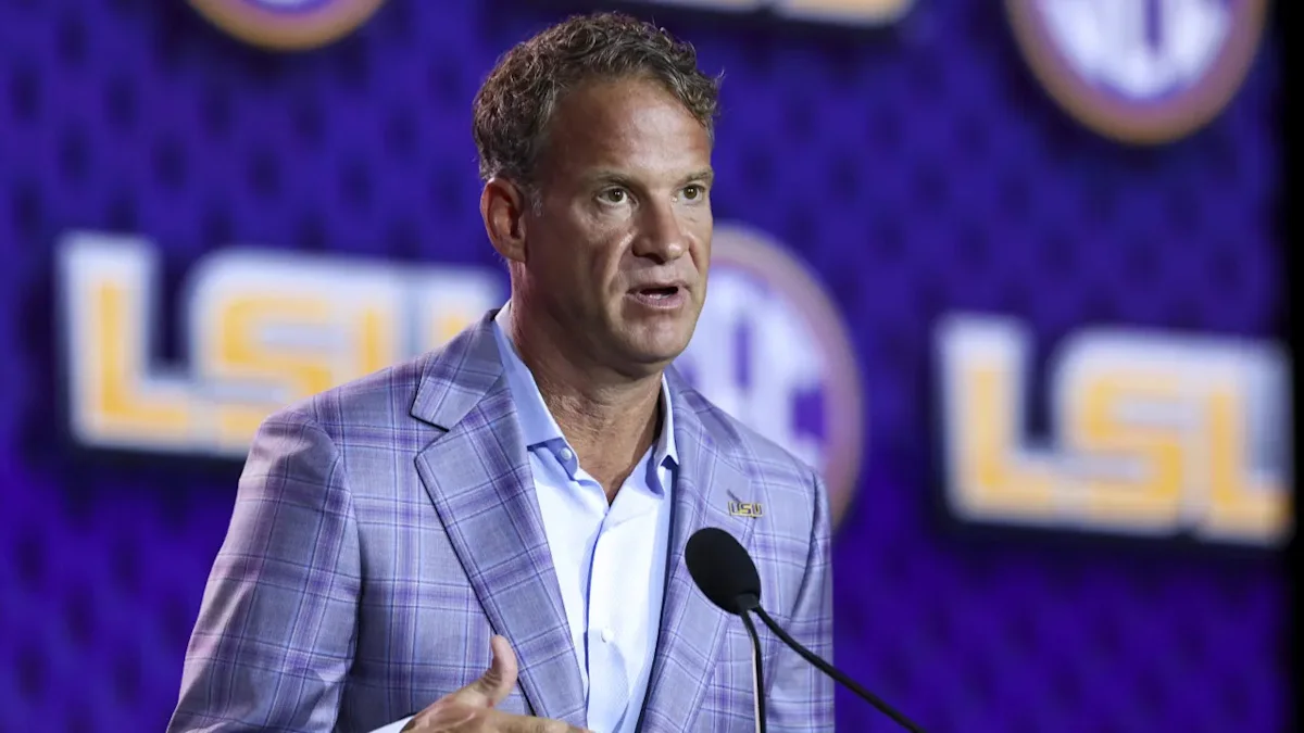 Man in a purple plaid jacket speaks at a microphone against a purple LSU backdrop
