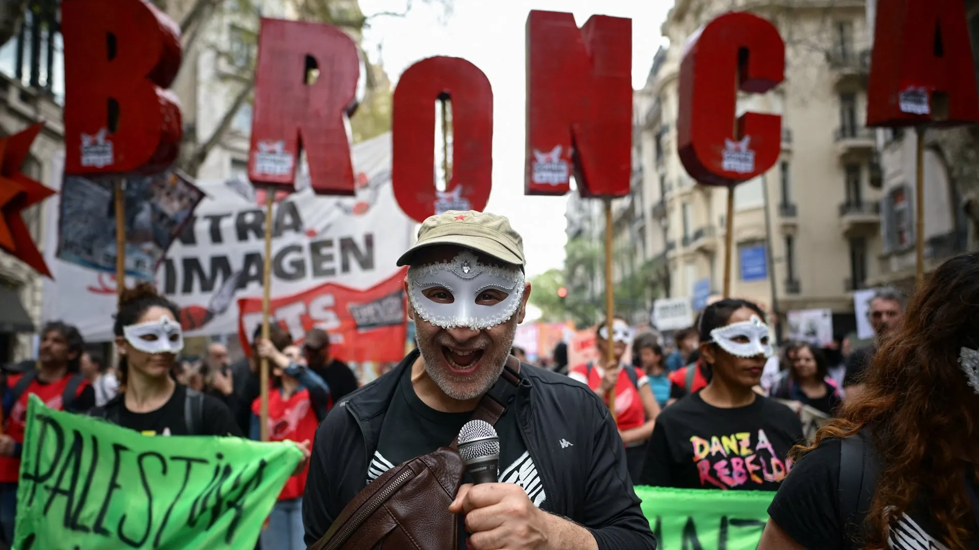Masked protester holding a microphone amid a crowded street demonstration with banners and large red letters
