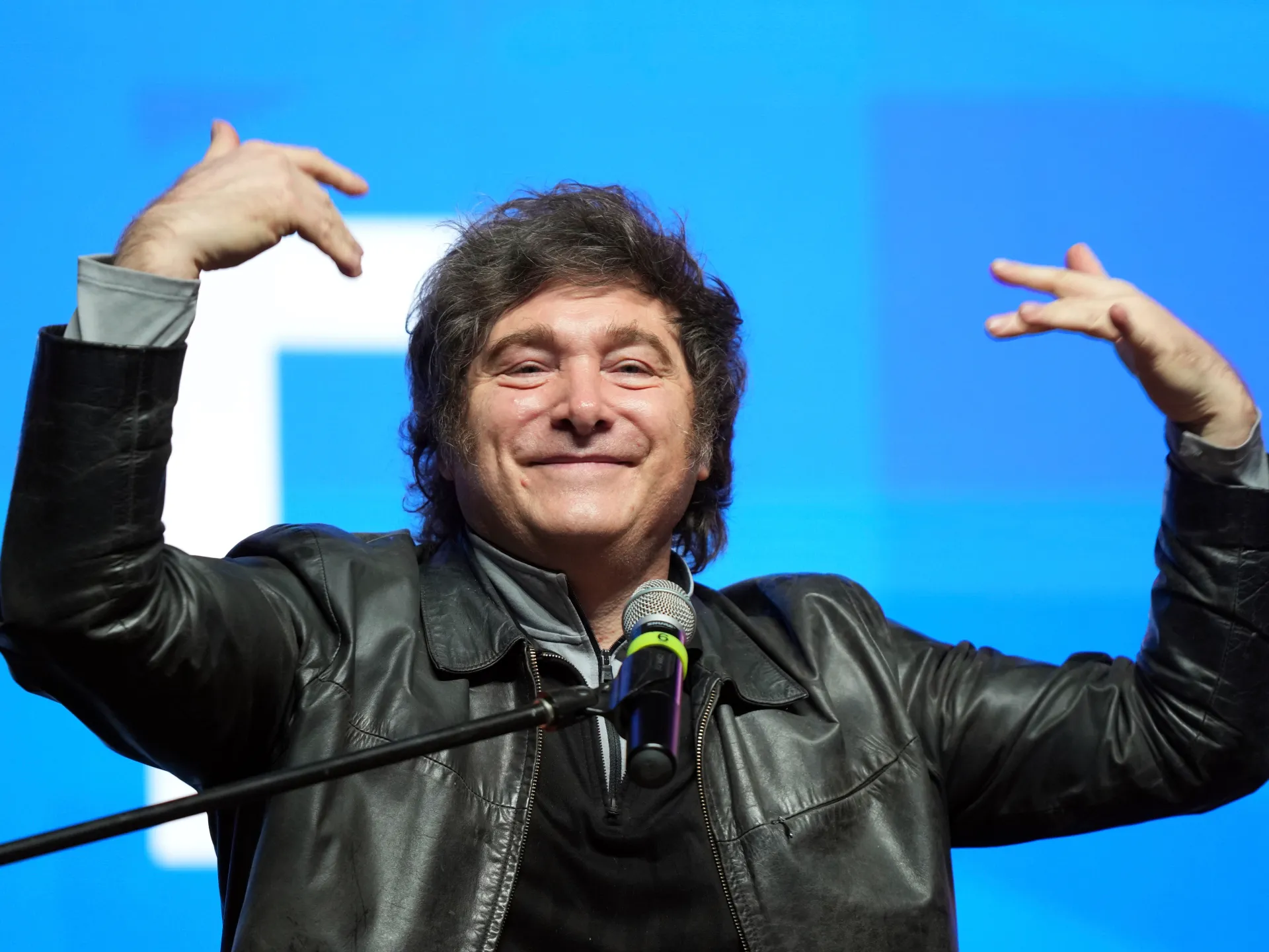 A man in a black leather jacket gestures with both hands beside a microphone against a blue backdrop.