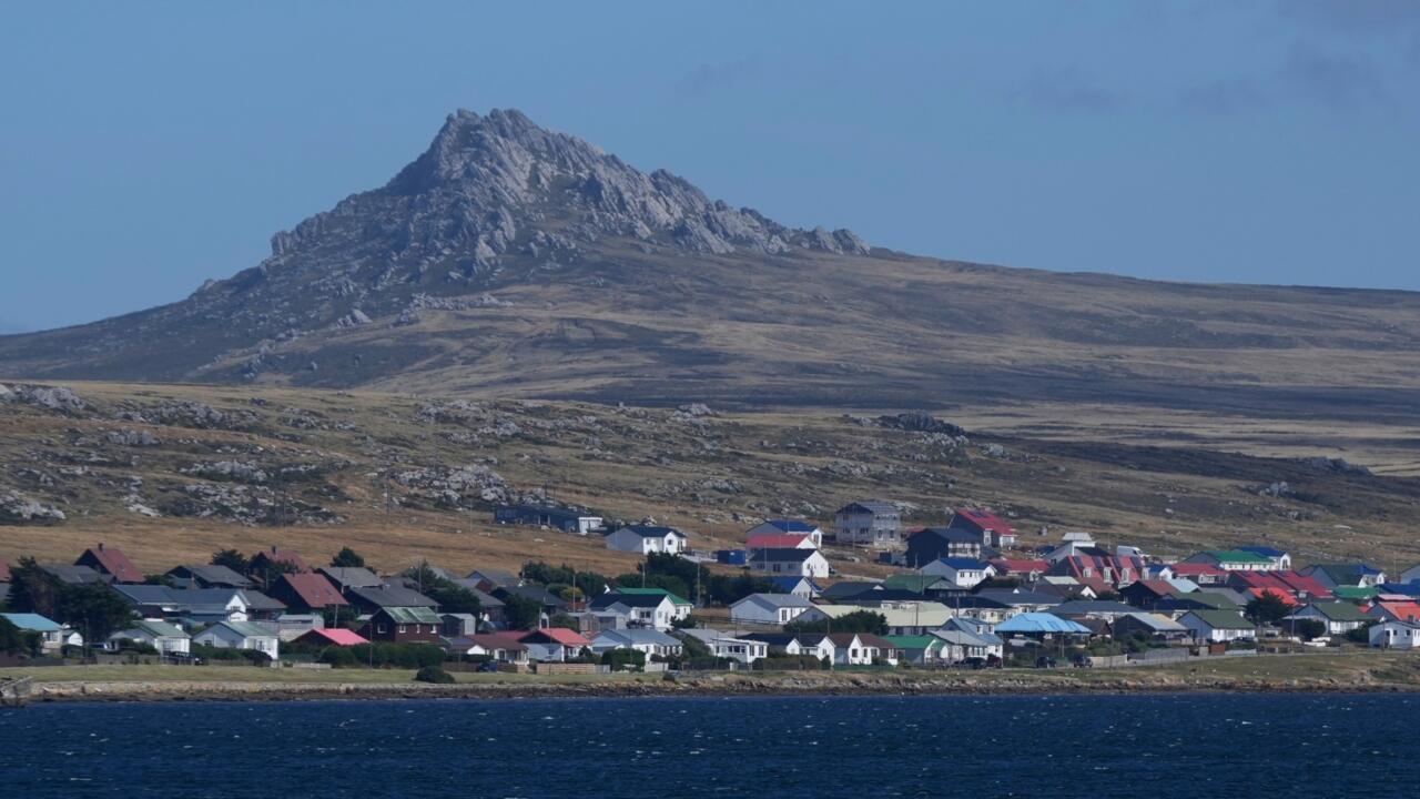 A coastal settlement with colorful rooftops sits beneath a rocky mountain beside dark blue water.
