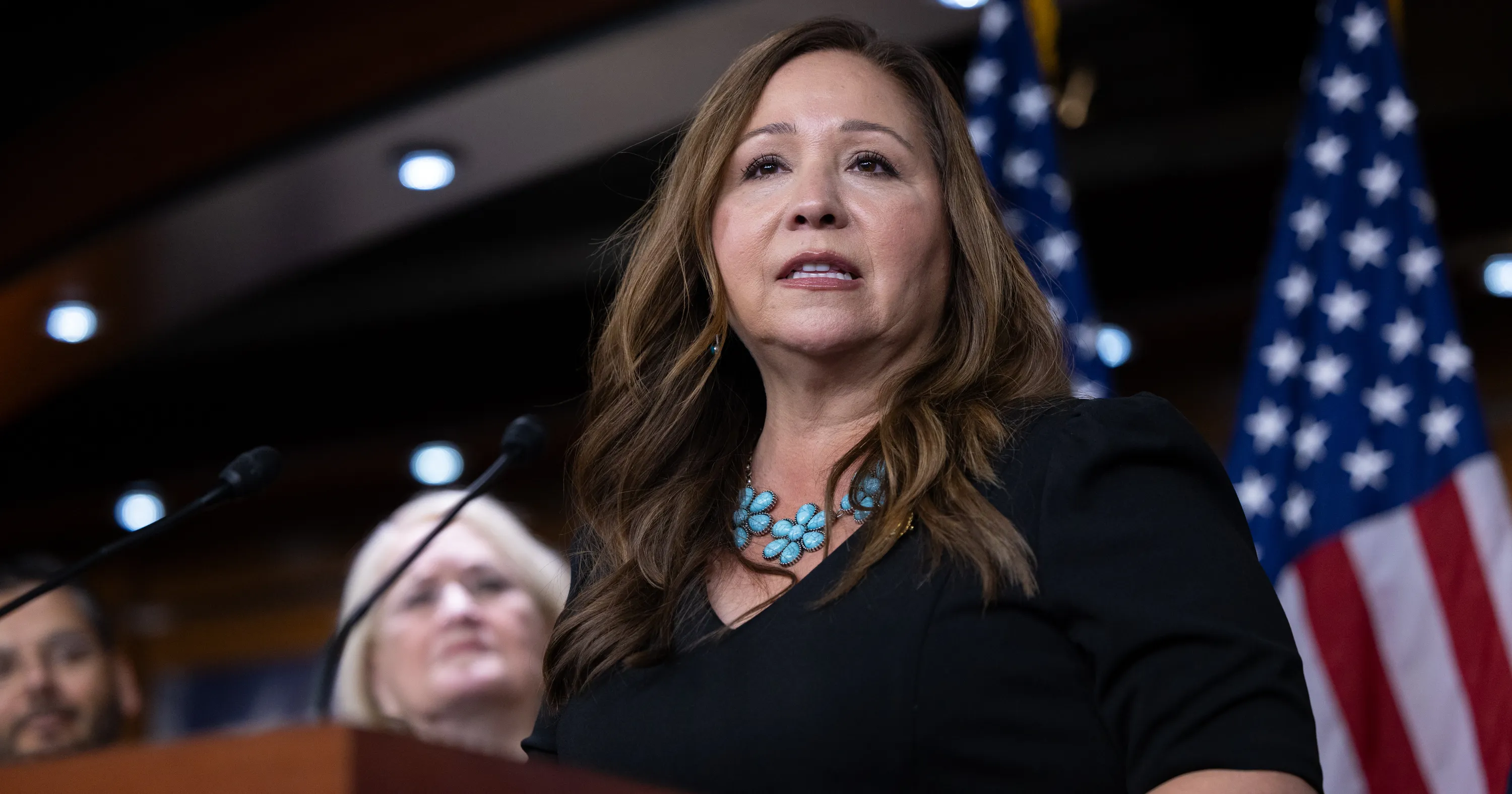 A woman speaks at a lectern with microphones, an audience, and American flags behind her.