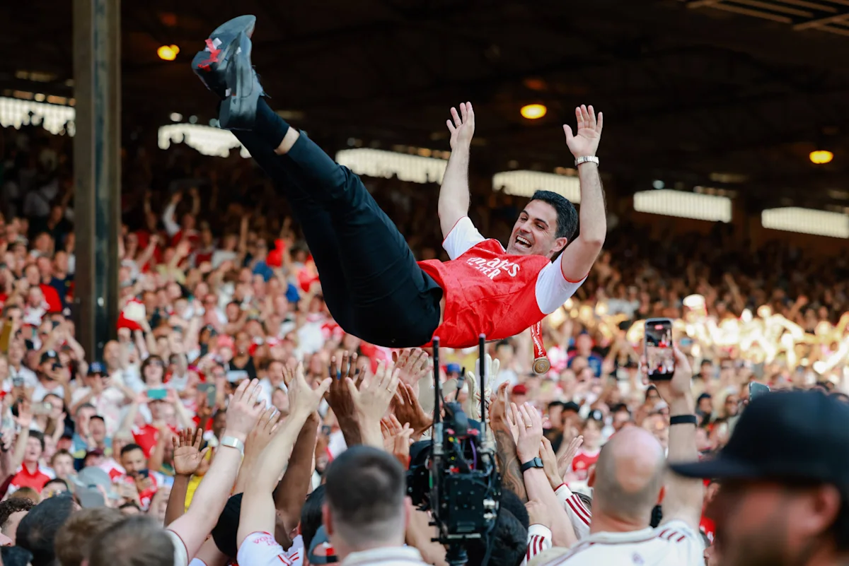 Footballer in red shirt crowdsurfs above cheering fans with arms raised during a celebration.