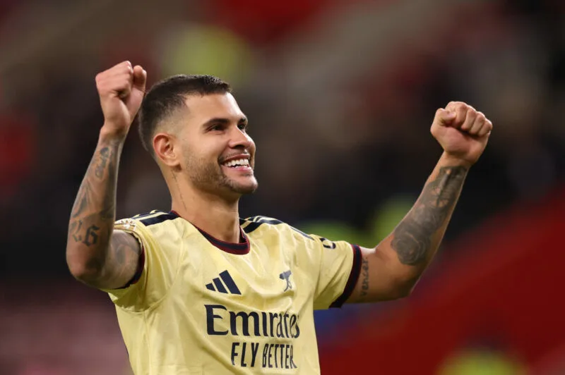 Arsenal footballer in a yellow jersey raises both tattooed arms and smiles.