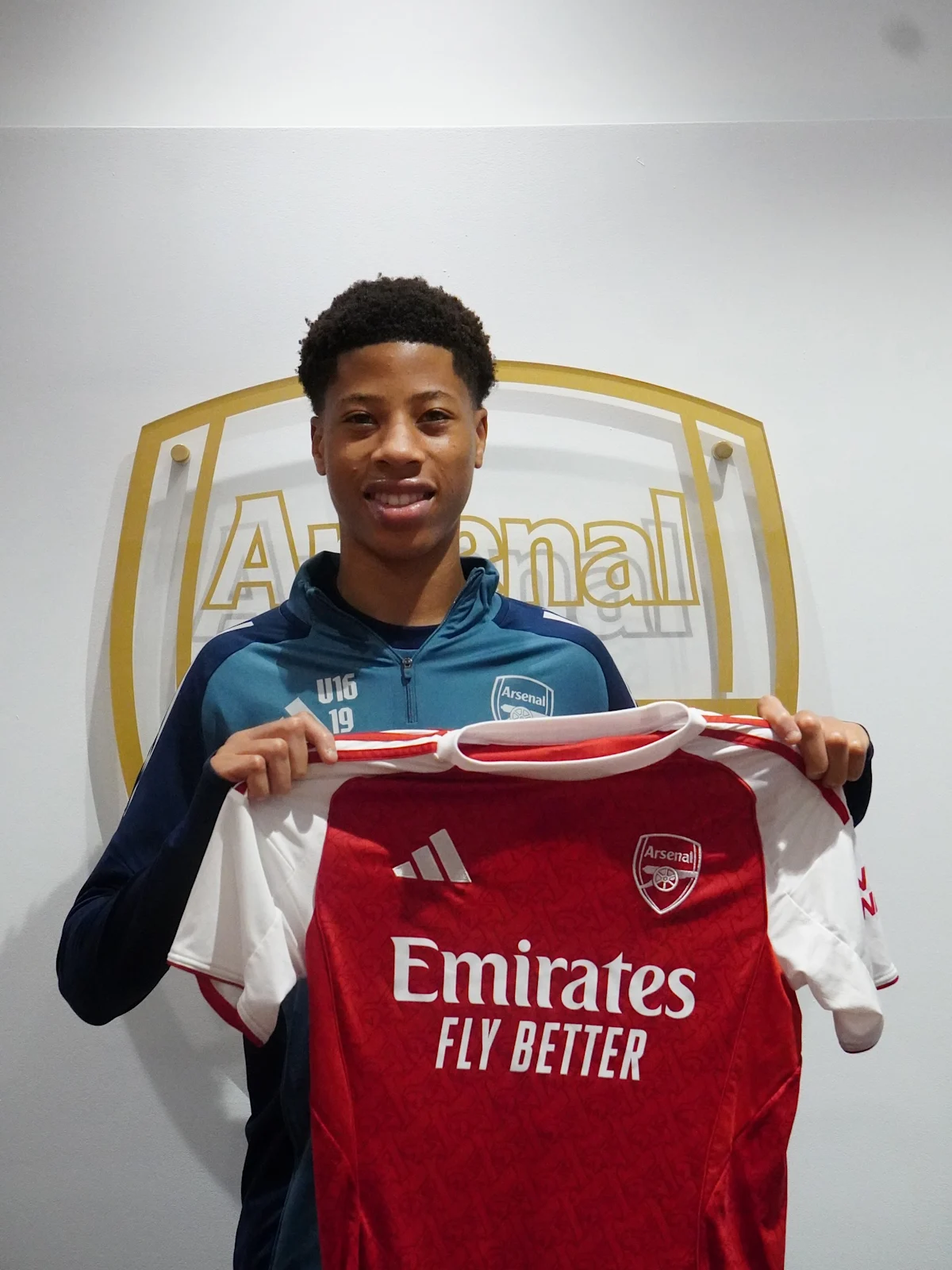Young Arsenal player holds a red-and-white football shirt in front of an Arsenal sign