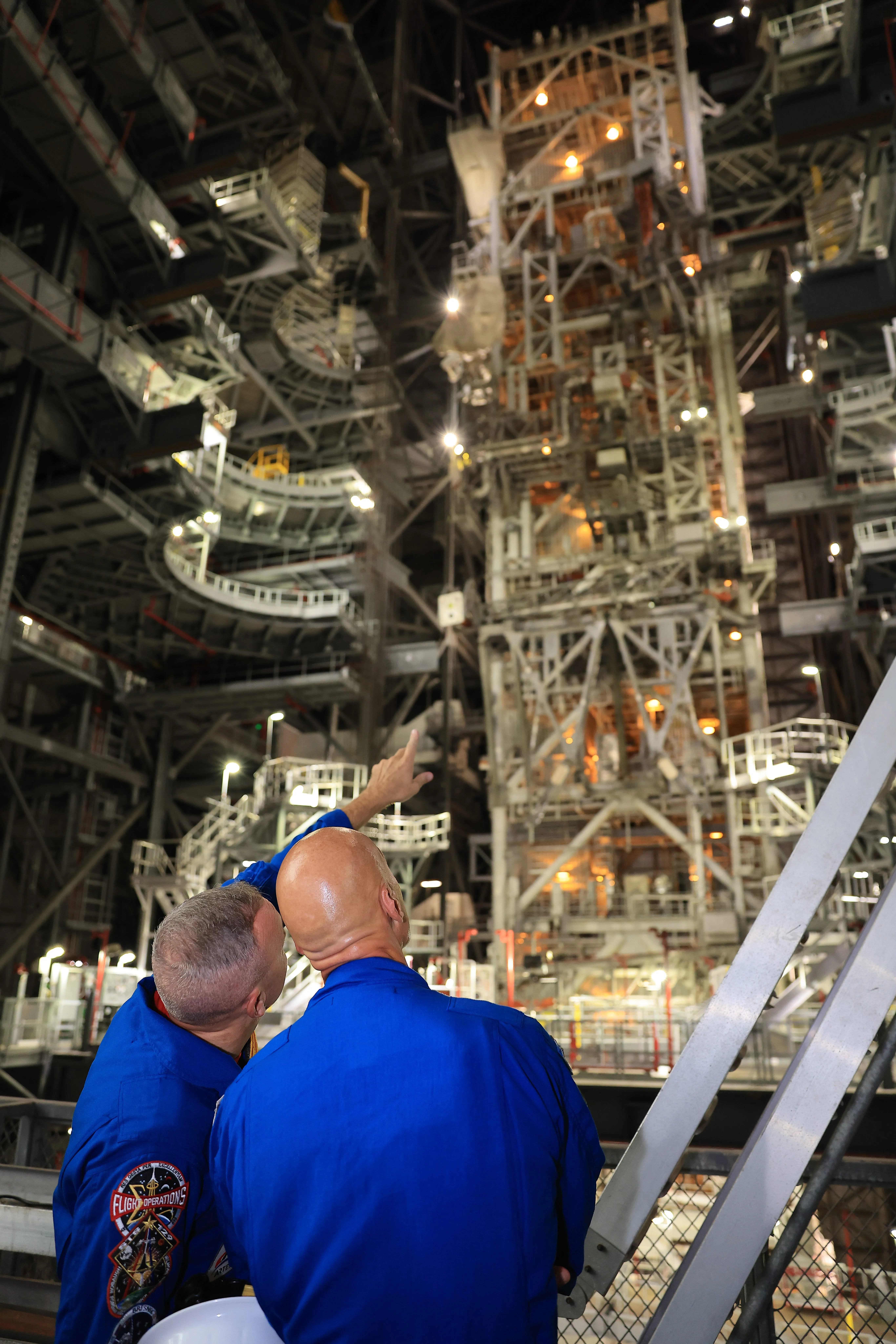 Two people in blue flight suits look up at industrial machinery inside a large assembly facility.